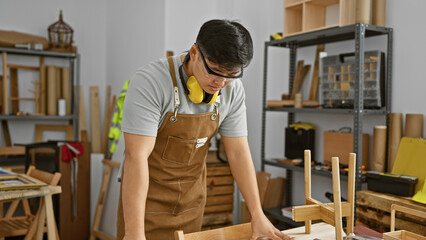 A young asian man examines wooden furniture intricately in a well-organized carpentry workshop.