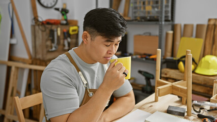 Fototapeta premium Young asian man enjoys a coffee break in a well-equipped carpentry workshop, surrounded by tools and wood.