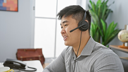 A cheerful young asian man wearing a headset in a modern office setting, engaged in customer...