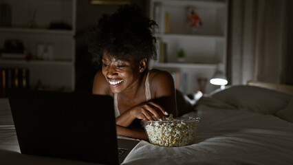 African american woman wearing lingerie watching movie on laptop eating popcorn at bedroom - obrazy, fototapety, plakaty