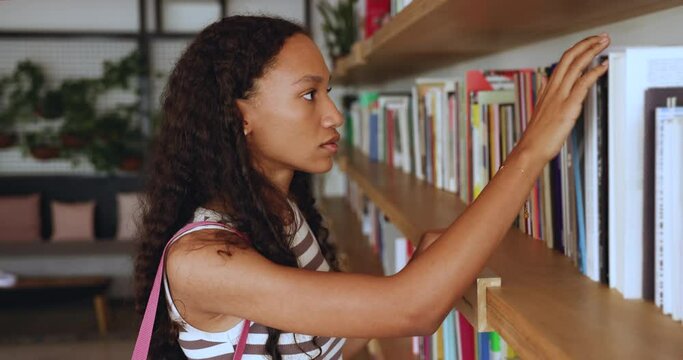 Close up portrait of happy young African teenager student girl standing near bookshelves in pubic or university library, choose book, smile, looking at camera. Studentship, new knowledge, education