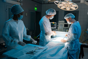 Remote view of skilled diverse surgeons and nurse wearing sterile gloves, surgical uniforms and masks processing surgical operation in dark operating room, of modern hospital emergency department.