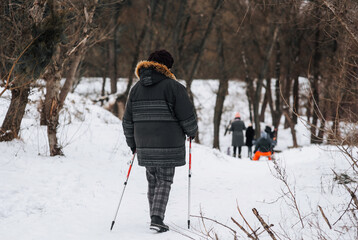 Fototapeta na wymiar An adult, elderly woman with aluminum hiking sticks walks along the road in winter on white snow on the street outdoors. Photography, healthy lifestyle concept.