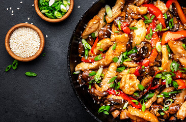 Hot stir fry chicken  slices with red paprika, mushrooms, chives and sesame seeds in frying pan. Black table background, top view