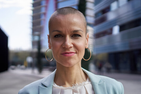 Close-up portrait of empowered middle-aged Caucasian business woman looking confident at camera. Formal attractive female posing elegant outdoors. Shaved head white person with successful expression