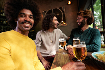 Happy portrait of an African american young guy with afro style smiling and looking at camera while enjoying time with friends in restaurant, drinking draft beer inside the bar. Friendly conversation.