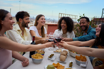 Fototapeta premium Laughing group diverse young friends enjoying lunch together outdoors. Cheerful people gathered toasting glass red wine on summer having fun celebrating birthday snack party on rooftop. Happy cheers