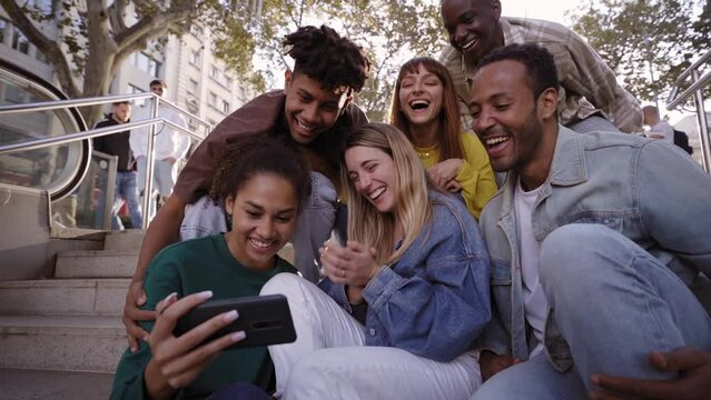 Diverse group of university friends sitting on stairs in street city. Happy students using and looking smartphones while smiling thrilled and excited. Chatting and commenting fun things in mobiles.