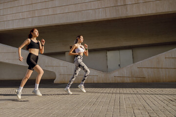 Two young women in sportswear are running on modern buildings background. Active lifestyle concept