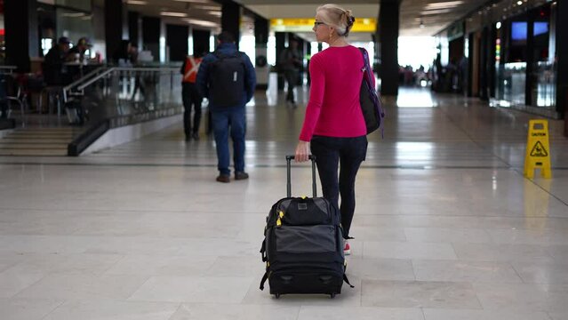 Pretty smiling mature woman walks through in airport wearing backpack and pulling luggage to her gate.