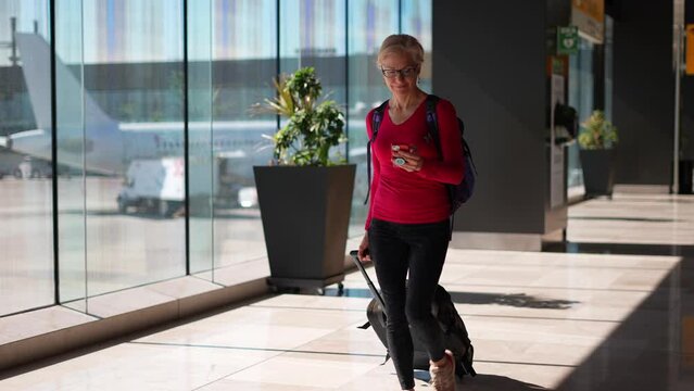 Pretty smiling mature woman walks through in airport wearing backpack and pulling luggage to her gate.
