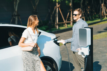 Young couple travel with EV electric car charging in green sustainable city outdoor garden in...