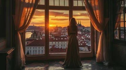 Tuinposter A graceful lady standing by a large window with a view of historic buildings in the city of Prague, Czech Republic in Europe. © Joyce