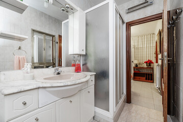A small bathroom with white porcelain toilets on glossy white wooden furniture, framed mirror on the wall