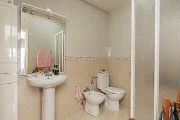 A bathroom with a white porcelain sink with a base of the same material under a frameless mirror with glass shelf and other matching toilets