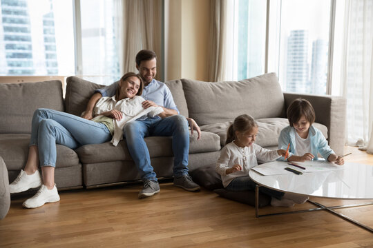 Young loving couple with preschoolers children spend time in living room, kids painting with pencils, parents relaxing on cozy couch, enjoy pastime together at modern home. Family leisure, hobby, rest