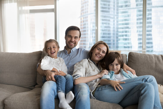 Young wife and husband posing for family photo sit on sofa with two little kids, smile look at camera, spend time together at modern apartment. Bank loan, new dwelling owners portrait, custody, love