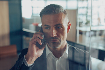 Handsome male manager in business casual clothes is talking phone with client in modern office 