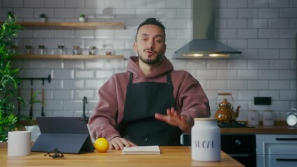 Vlogger man in an apron sit in kitchen talking to camera, recording video about making lemonade at domestic home. Online broadcast, man leads food blog or course - Powered by Adobe