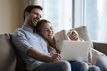 Happy spouses enjoy time together, relaxing on couch with laptop, laugh, watch funny videos...