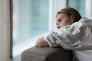 Lonely young woman lies on sofa, looking out window, having melancholic mood, suffer of unrequited...