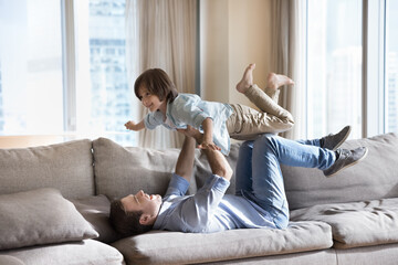 Young father enjoy playtime with son, lying on sofa lifts kid on arms, happy boy pretend fly in...