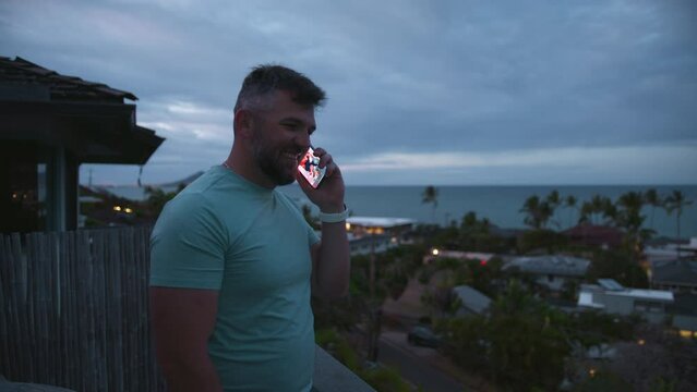 Bearded businessman free lancer working remotely answer call. Positive healthy guy standing on balcony with tropical island views on overcast day. Athletic smiling guy talking outside using smartphone