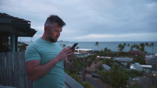 Mature handsome guy standing on balcony with tropical island views on overcast evening. Athletic guy looking at smartphone screen. Bearded businessman free lancer working remotely answering emails 4K