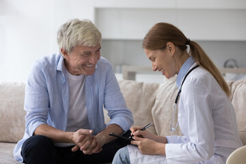 Positive elderly man consulting young doctor woman at home, speaking to medical specialist, smiling, laughing, feeling healthy. Practitioner visiting senior male patient in apartment, writing notes