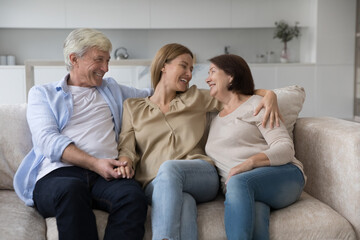 Positive caring senior parents and loving adult daughter woman enjoying leisure, closeness, sitting on couch at home together, hugging, smiling, discussing family news, laughing