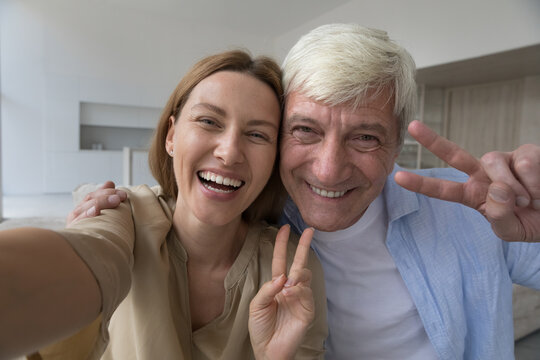 Funny family self portrait of cheerful senior dad and pretty adult daughter child holding device in hand, smiling at camera, laughing, taking home selfie making video call, showing victory fingers