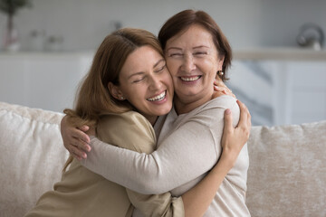 Cheerful pretty mature mom embracing, cuddling daughter on home couch, laughing, hugging adult...