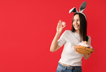 Pretty young woman with bunny ears and Easter basket on red background