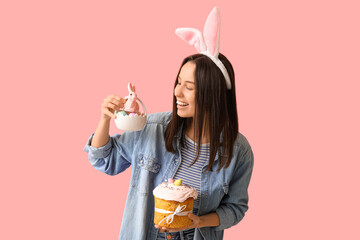 Pretty young woman with bunny ears, Easter cake and basket on pink background