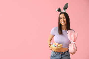 Pretty young woman with bunny ears, Easter basket and gift on pink background