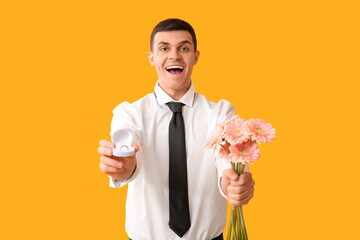 Handsome man with flowers and engagement ring on yellow background