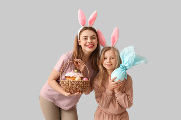 Cute little girl with gift egg and her mother in bunny ears holding basket with Easter cake on grey background