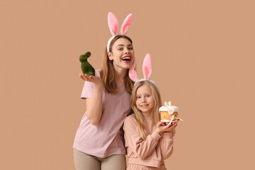 Cute little girl with Easter cake and her mother in bunny ears holding rabbit on brown background