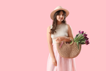 Pretty young woman holding wicker bag with bouquet of beautiful purple tulips on pink background