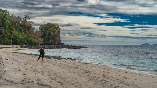 A girl stands on a sandy beach, taking pictures of a picturesque rock. The view from the back. Footprints in the sand. A calm turquoise ocean. Clouds in the blue sky. Madagascar. Nosy Tanikely 