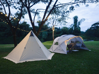 Outdoor camping tent with tarp on grass courtyard and warm night light under dark blue sky twilight time