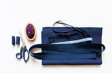 Materials for handmade sewing bag for shopping: thick blue fabric, sling for handles, braid, scissors, thread, needle bed on white work table. Flat lay, copy space, top view