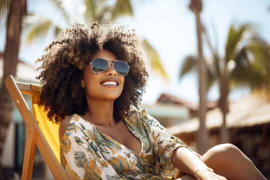 Portrait of happy young black woman relaxing on wooden deck chair at tropical beach while looking at camera wearing spectacles. Smiling african american girl with fashion sunglasses enjoying vacation.