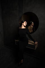 Sexy woman in a black dress posing near a mirror in a dark room. Fashion shooting concept