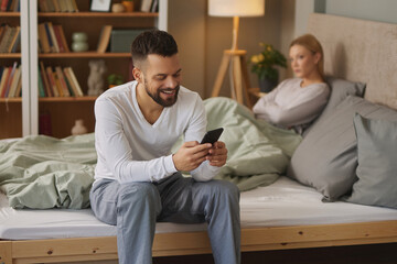 Man with smartphone in bedroom, upset woman in the background