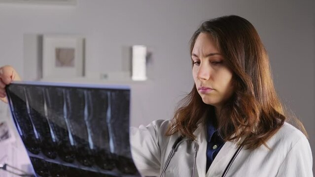 A young female doctor in a white coat holds an X-ray of the bones of the spine, examines it, then looks at the camera. Medicine and treatment of protrusion and fractures