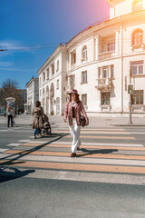 Woman city road crossing. Stylish woman in a hat crosses the road at a pedestrian crossing in the city. Dressed in white trousers and a jacket with a bag in her hands.