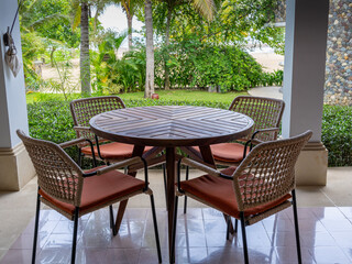 Obraz premium A coffee table set with one circular wooden table and four simple armchairs set up at a clean terrace overseeing a tidy garden by the beach and pool.