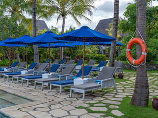 Rows of poolside and outdoor lounge chairs with blue and white color theme and placed strategically surrounding the pool and by the beach sand near a beach bar.