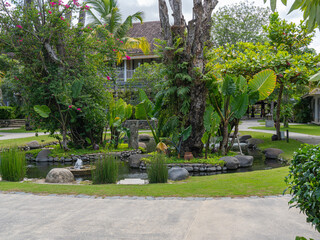 A circular small green garden with a fish pond at the middle, green trees, well maintained green grass, and a concrete pathway.